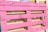 A wooden pallet painted in bright pink, with natural wood color visible in the background. The pallet is leaning against other unpainted pallets, showing a contrast between the vivid pink and the neutral tones of wood.