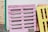 A stack of wooden pallets painted in various colors, including pink and yellow, leaning against a white wall. They are outside on a gravel surface with a portion of a wooden fence visible on the left.