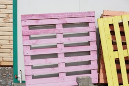A stack of wooden pallets painted in various colors, including pink and yellow, leaning against a white wall. They are outside on a gravel surface with a portion of a wooden fence visible on the left.