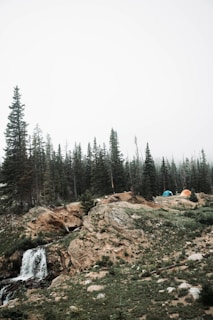 A mountainous landscape featuring a small waterfall cascading over rocks surrounded by lush greenery and pine trees. Two colorful tents, one blue and one orange, are set up on a rocky plateau amidst the misty atmosphere.