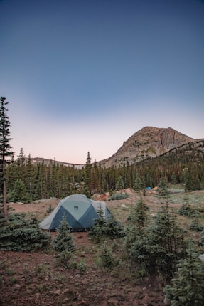 A camping tent is set up in a forested area with numerous pine trees. In the background, a rugged mountain peak rises against a clear sky transitioning from light near the horizon to deep blue overhead. The landscape is rugged with patches of grass and scattered small plants.