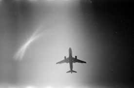 A black and white image of an airplane flying high in the sky. The aircraft is silhouetted against a gradient background with light coming from one side, creating an ethereal and dramatic effect. The composition focuses on the aircraft from a bottom-up perspective.