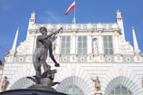 A detailed, ornate fountain featuring a large statue of a male figure, possibly Neptune, holding a trident and standing over a mythological creature in front of an elegant, historical building. The building has intricate architectural details, large windows, and is topped with a red and white flag.