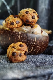 A wooden bowl filled with freshly baked chocolate chip cookies sits on a patterned fabric surface. One cookie is placed in front of the bowl, displaying its golden-brown texture and rich chocolate chips. The background is softly blurred, emphasizing the rustic and homemade appeal of the cookies.