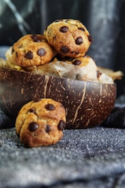 A wooden bowl filled with freshly baked chocolate chip cookies sits on a patterned fabric surface. One cookie is placed in front of the bowl, displaying its golden-brown texture and rich chocolate chips. The background is softly blurred, emphasizing the rustic and homemade appeal of the cookies.