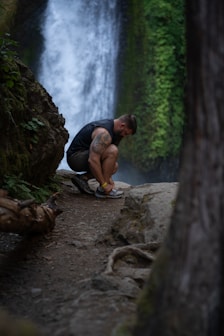 a man squatting on a rock in front of a waterfall