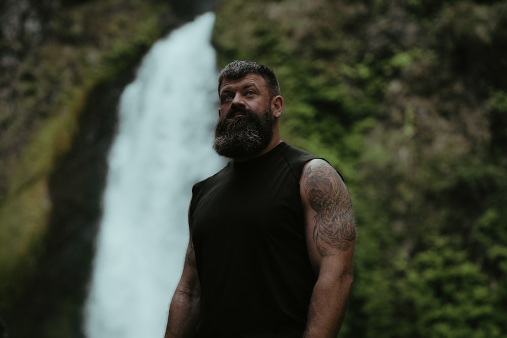 um homem com barba em pé em frente a uma cachoeira