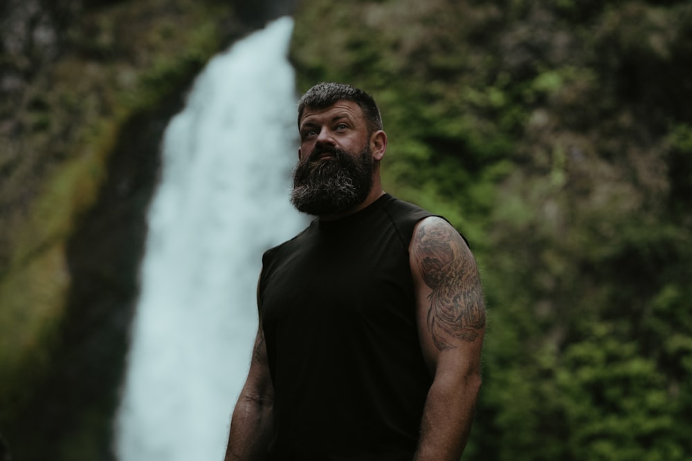 a man with a beard standing in front of a waterfall