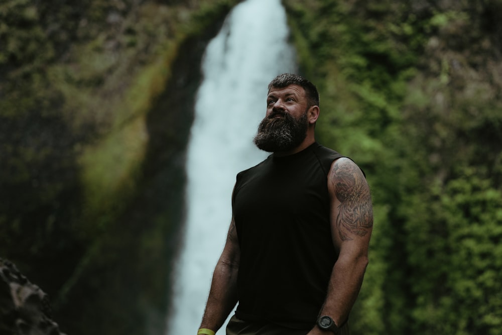 um homem com barba em pé em frente a uma cachoeira