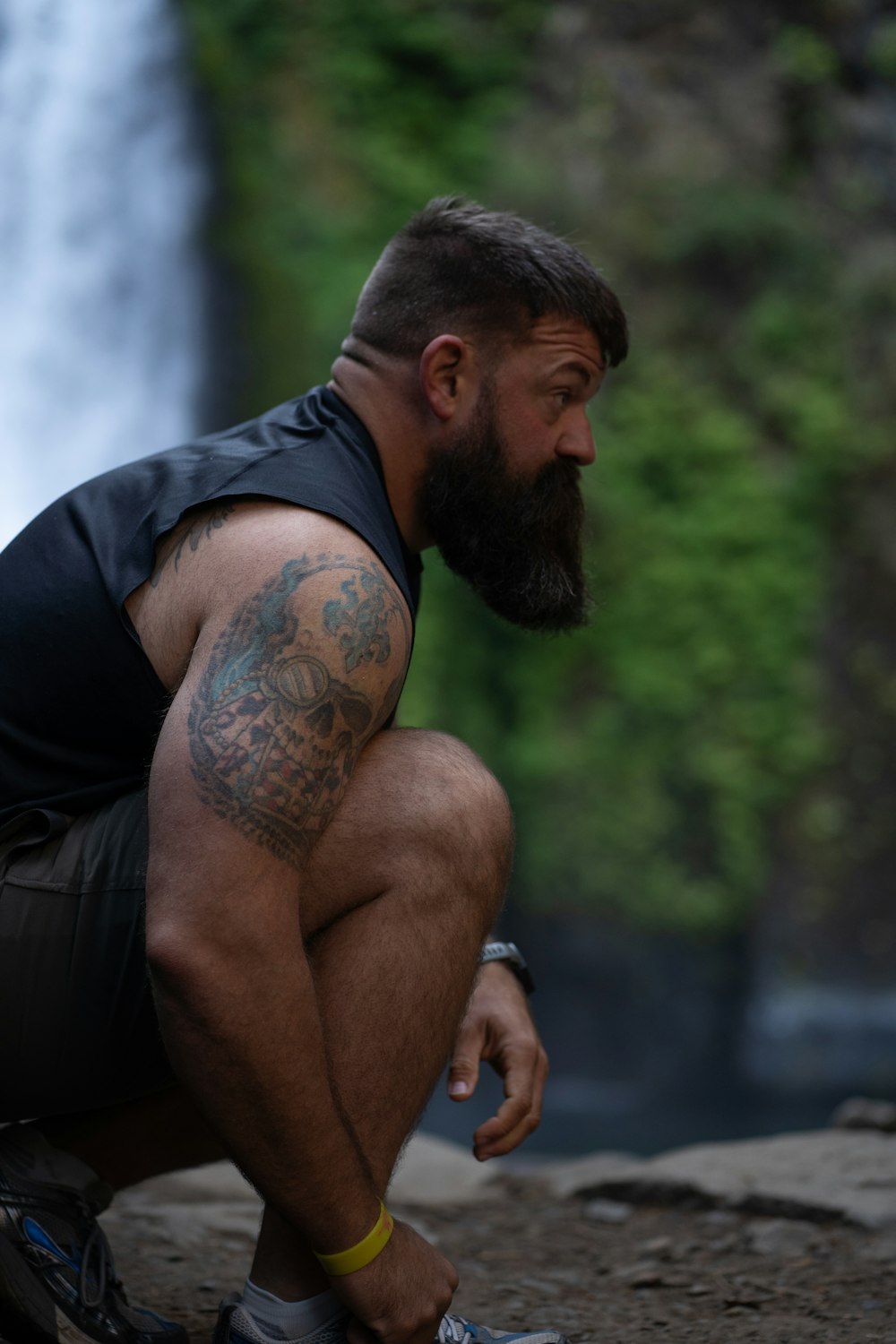 a man with a beard squatting in front of a waterfall