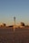A beach scene during sunset with golden sand and a tall pole with a circular element on top in the foreground. In the background, there are several wind turbines and modern buildings, including a cylindrical structure. A few people are walking or standing on the sand.