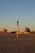 A beach scene during sunset with golden sand and a tall pole with a circular element on top in the foreground. In the background, there are several wind turbines and modern buildings, including a cylindrical structure. A few people are walking or standing on the sand.