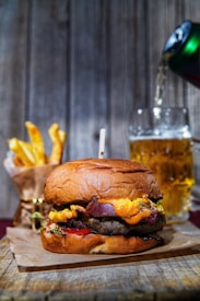 A juicy burger with melted cheese, crispy bacon, and fresh vegetables sits on a wooden board, garnished with a delicate paper wrap. Beside it, a bouquet of golden French fries in a paper holder adds to the appetizing display. A frothy beer is being poured from a green can into a clear mug in the background.
