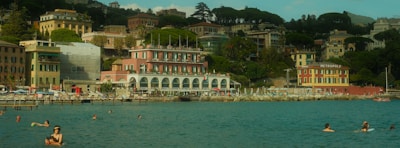 A picturesque coastal scene featuring swimmers enjoying the calm, blue water. In the backdrop, colorful buildings with a mix of architectural styles are nestled among lush green foliage. A hotel named Metropole is prominently visible, and people are relaxing by the waterfront, which is lined with lounge chairs and umbrellas.