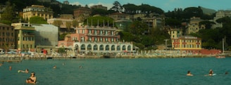 A picturesque coastal scene featuring swimmers enjoying the calm, blue water. In the backdrop, colorful buildings with a mix of architectural styles are nestled among lush green foliage. A hotel named Metropole is prominently visible, and people are relaxing by the waterfront, which is lined with lounge chairs and umbrellas.