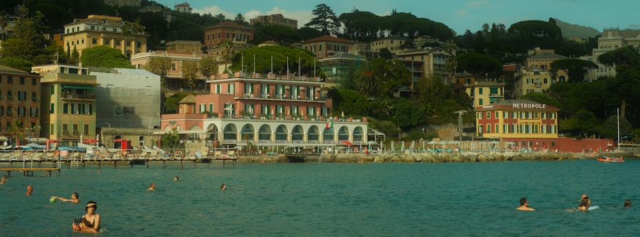 A picturesque coastal scene featuring swimmers enjoying the calm, blue water. In the backdrop, colorful buildings with a mix of architectural styles are nestled among lush green foliage. A hotel named Metropole is prominently visible, and people are relaxing by the waterfront, which is lined with lounge chairs and umbrellas.