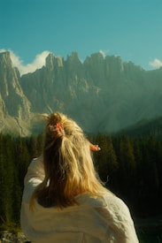 A person with blonde hair wearing a white shirt is standing in a forested area, gazing at a range of rugged mountains under a clear blue sky. The scene conveys a sense of wonder and connection with nature.