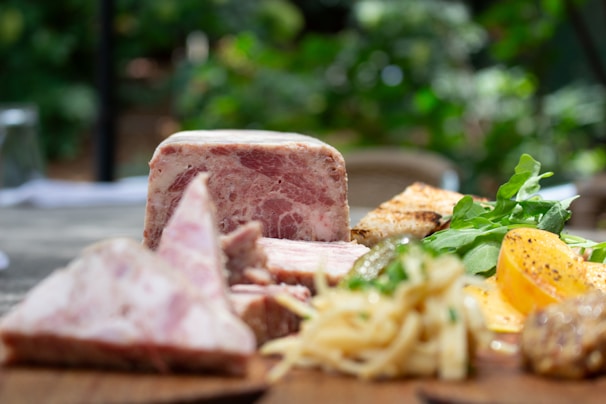 A platter of charcuterie and fresh greens is arranged on a wooden surface. Slices of cured meat and pâté are paired with what appears to be a side of toast, arugula, and sliced tomatoes. There are also some pickled items and a portion of shredded cheese or pasta.
