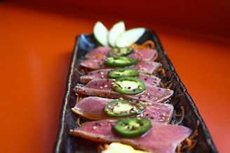 Sliced seared tuna arranged on a rectangular black plate, garnished with jalapeno slices and sprinkled with sesame seeds. Thin apple slices are placed at the end of the plate, all set against a warm orange background.