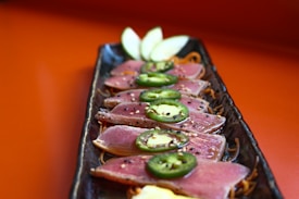 Sliced seared tuna arranged on a rectangular black plate, garnished with jalapeno slices and sprinkled with sesame seeds. Thin apple slices are placed at the end of the plate, all set against a warm orange background.