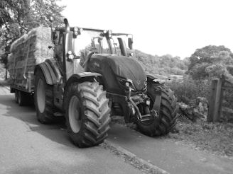 A large tractor pulling a trailer loaded with bales of hay is parked on the side of a rural road. The scene is surrounded by dense trees and foliage, suggesting a countryside setting.