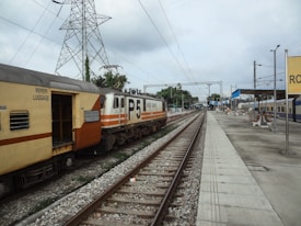 A railway platform with an orange and cream-colored train on the tracks, power lines overhead, and a few people in the distance. A signboard is visible on the right.
