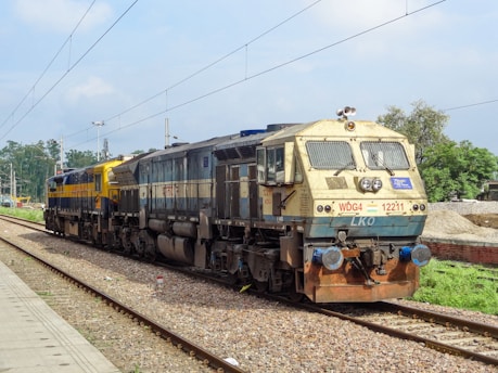A locomotive train is seen on a railway track surrounded by greenery. The engine is marked with the code WDG4 12211 and appears to be stationary. Overhead power lines and trees are visible in the background under a partly cloudy sky.