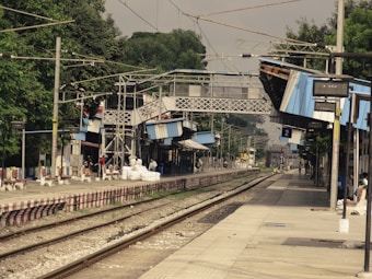 A railway station with multiple tracks and platforms is depicted, featuring a pedestrian overpass connecting the platforms. Several people are standing and waiting, with some packages and goods placed on the platform. The station has blue canopies overhead and lush green trees in the background.