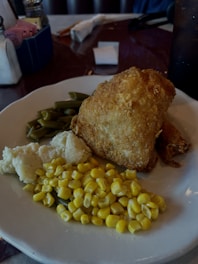A plate with breaded fried chicken, cooked green beans, mashed potatoes, and corn. The table setting includes a dark drink in a glass and a small basket with sugar packets and condiments in the background.