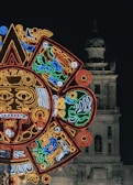 A vibrant, illuminated design resembling an Aztec or Mayan calendar is prominently displayed against the backdrop of an old, historical building. The building, which appears to be a cathedral, features a bell tower and intricate stonework.