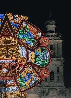 A vibrant, illuminated design resembling an Aztec or Mayan calendar is prominently displayed against the backdrop of an old, historical building. The building, which appears to be a cathedral, features a bell tower and intricate stonework.