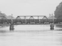 A black and white image capturing a metal bridge spanning across a calm river. The bridge features a train with cargo or freight cars traveling across, supported by piers submerged in the water. Trees and some infrastructure can be seen in the distance on both sides of the river.