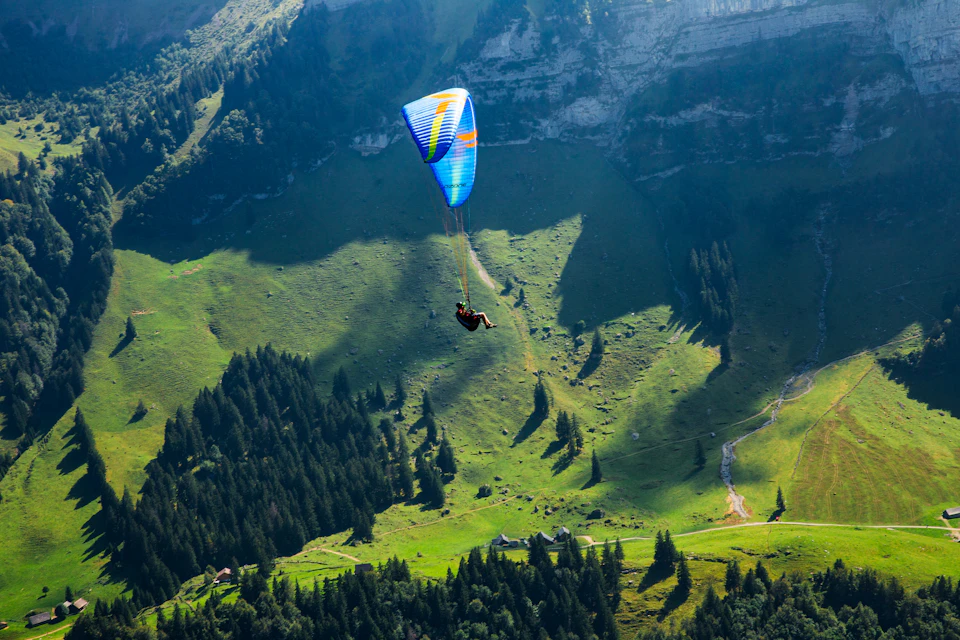 The image depicts a person paragliding over a verdant mountain range, with their colorful parachute billowing against the clear blue sky. The paraglider is positioned near the top left corner of the frame and appears to be in mid-flight, soaring above the lush green landscape below.

In the background, there are several trees dotting the mountainside, adding depth and texture to the scene. The image also contains a few buildings scattered across the mountain range, possibly serving as accommodations or base camps for other adventurers.