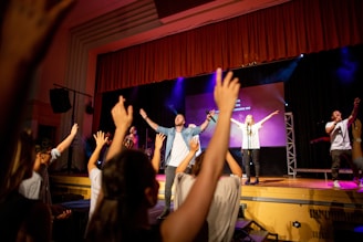 A group of people is on a stage in front of a cheering audience. The individuals on stage are holding microphones and engaging the crowd with their arms raised. The background features stage lights and a large screen displaying text. Audience members also have their hands raised, participating enthusiastically.