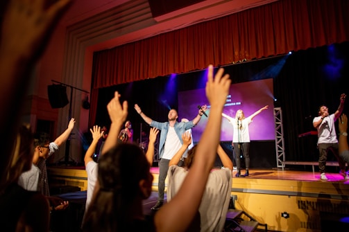 A group of people is on a stage in front of a cheering audience. The individuals on stage are holding microphones and engaging the crowd with their arms raised. The background features stage lights and a large screen displaying text. Audience members also have their hands raised, participating enthusiastically.