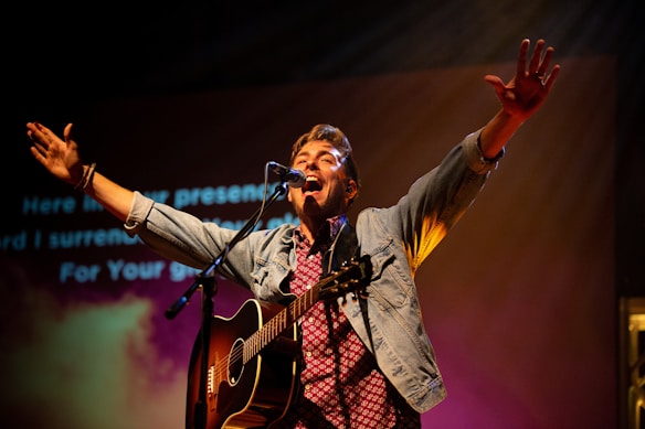 A musician passionately performs on stage with a guitar, arms wide open, and a microphone in front of him. The backdrop features projected text and colorful lights, creating a dynamic and engaging concert atmosphere.