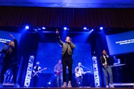 A live performance on stage with musicians and singers. Several people are holding microphones and instruments, including guitars. The stage is illuminated with blue and white lights, and a screen in the background displays text.