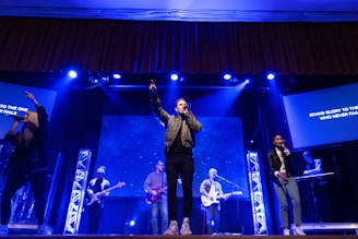 A live performance on stage with musicians and singers. Several people are holding microphones and instruments, including guitars. The stage is illuminated with blue and white lights, and a screen in the background displays text.
