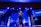 A live performance on stage with musicians and singers. Several people are holding microphones and instruments, including guitars. The stage is illuminated with blue and white lights, and a screen in the background displays text.