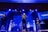 A live performance on stage with musicians and singers. Several people are holding microphones and instruments, including guitars. The stage is illuminated with blue and white lights, and a screen in the background displays text.