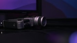 A vintage film camera is placed on a dark reflective surface with a soft focus background displaying abstract colors on a monitor.
