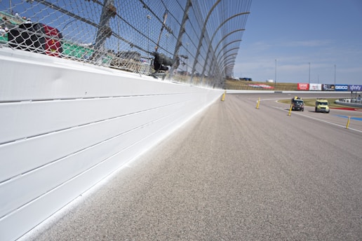 A racetrack is depicted with a high boundary wall and a protective chain-linked fence that runs alongside the track. There are two cars on the track, and various safety barriers and poles are placed along the road. In the background, there are advertisements and a clear blue sky.