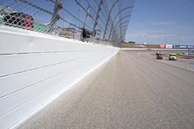 A racetrack is depicted with a high boundary wall and a protective chain-linked fence that runs alongside the track. There are two cars on the track, and various safety barriers and poles are placed along the road. In the background, there are advertisements and a clear blue sky.
