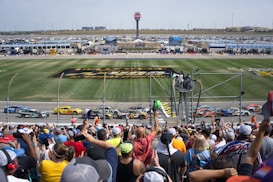 A crowd of enthusiastic spectators watches a car race at a large race track. Several brightly colored race cars are speeding past the spectators, with some fans waving and taking pictures. In the background, there are racing facilities, including stands, a digital screen, and a field with racing logos.