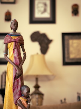 A tall, intricately carved wooden statue of a woman in traditional African attire, adorned with colorful beads and standing beside a smaller statue of a child. The background includes framed art and masks on the wall, along with a softly lit lamp creating a warm ambiance.