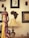 A tall, intricately carved wooden statue of a woman in traditional African attire, adorned with colorful beads and standing beside a smaller statue of a child. The background includes framed art and masks on the wall, along with a softly lit lamp creating a warm ambiance.