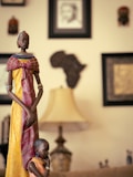 A tall, intricately carved wooden statue of a woman in traditional African attire, adorned with colorful beads and standing beside a smaller statue of a child. The background includes framed art and masks on the wall, along with a softly lit lamp creating a warm ambiance.