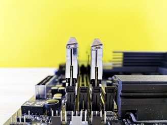 A close-up view of a computer motherboard with two RAM sticks installed vertically. The background is a bright yellow, and the board features various components, such as capacitors and heat sinks, with a mix of black, silver, and grey elements.