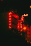 A building with red neon signs displaying the words 'HOTEL' and 'PERSIA' against a dark nighttime backdrop. The signs are both in English and a script which appears to be Persian. The building exterior has a modern design, prominently lit by the red glow from the signage.