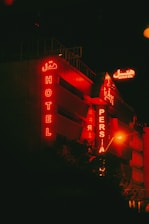 A building with red neon signs displaying the words 'HOTEL' and 'PERSIA' against a dark nighttime backdrop. The signs are both in English and a script which appears to be Persian. The building exterior has a modern design, prominently lit by the red glow from the signage.